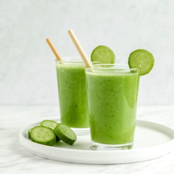 Two glasses of cucumber pineapple smoothie garnished with sliced cucumber with a white background.