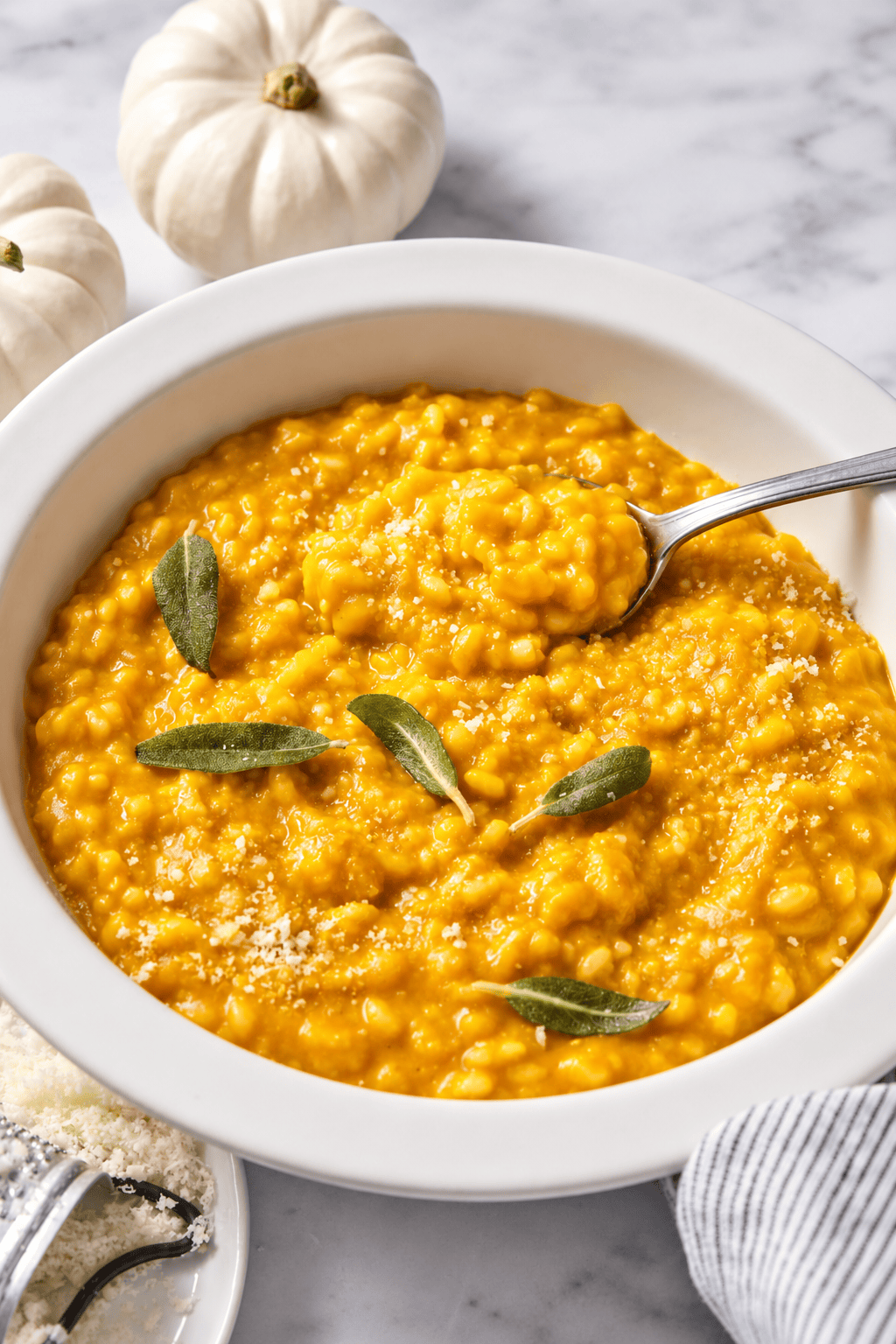 A white bowl of creamy vegan pumpkin risotto garnished with sage leaves, with a spoon, sits on a marble surface near small white pumpkins.