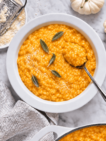 A bowl of creamy vegan pumpkin risotto topped with sage leaves and grated cheese, with a spoon in the dish. A grater, cheese, pumpkins, and a striped cloth are nearby on a marble surface.