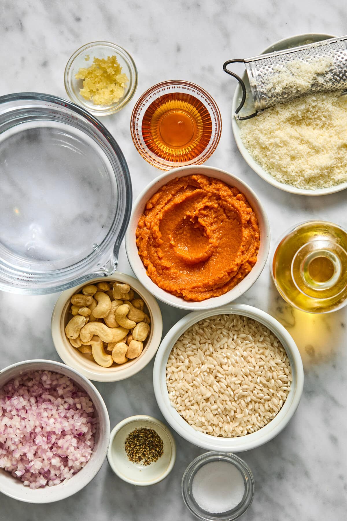 Bowls of ingredients on a marble surface include water, grated cheese, minced garlic, oil, red sauce, cashews, rice, chopped shallots, ground pepper, and salt-everything you need to create a flavorful vegan pumpkin risotto.