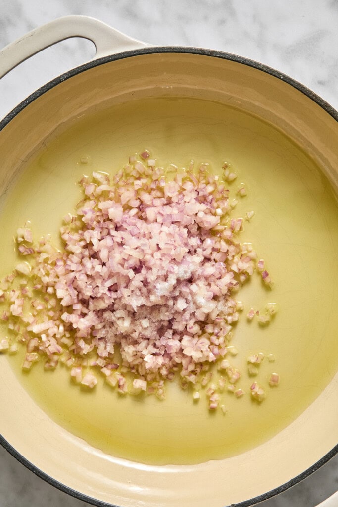 Chopped shallots sprinkled with salt in a large pot filled with olive oil, ready to be cooked on a marble countertop as the savory base for a vegan pumpkin risotto.