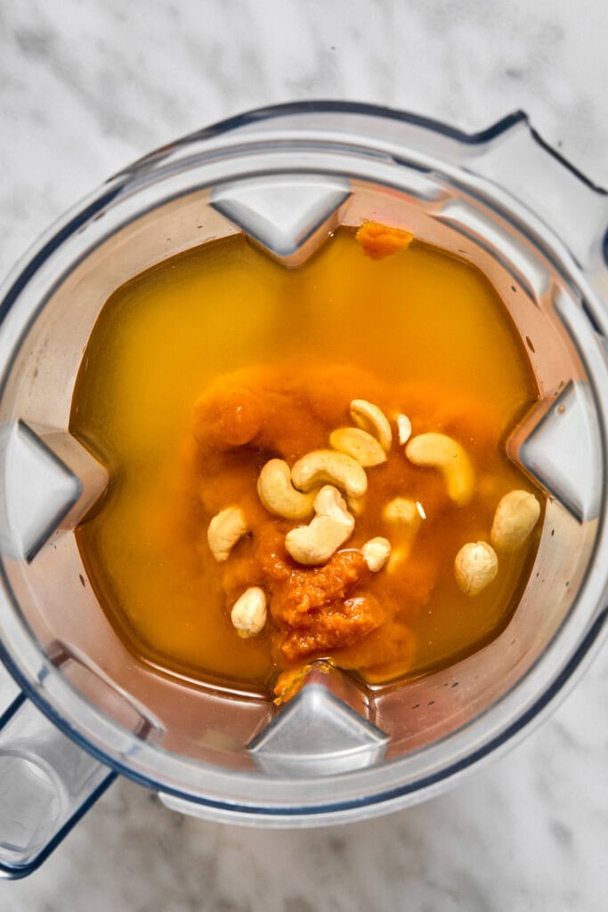 A blender contains orange liquid, pumpkin puree, and raw cashews, ready to be blended for a creamy vegan pumpkin risotto on a marble countertop.