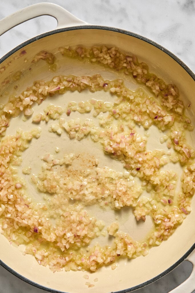Diced onions sautéing in oil in a cream-colored Dutch oven on a light marble surface, forming the aromatic base for a vegan pumpkin risotto.