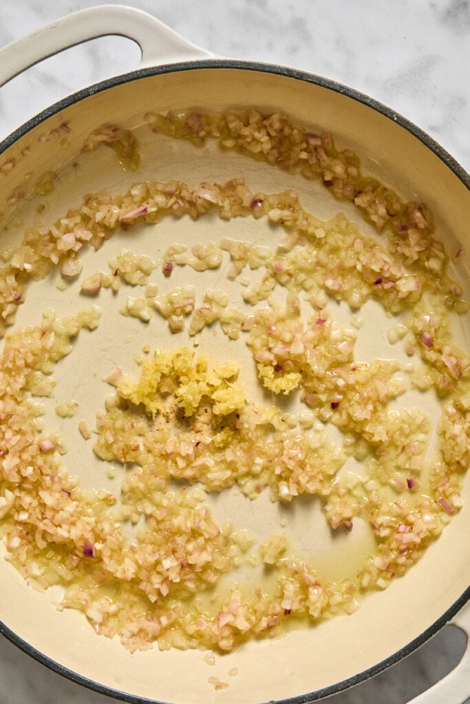 Diced onions and minced garlic sautéing in olive oil in a large cream-colored pot on a marble countertop, forming the savory base for a comforting vegan pumpkin risotto.
