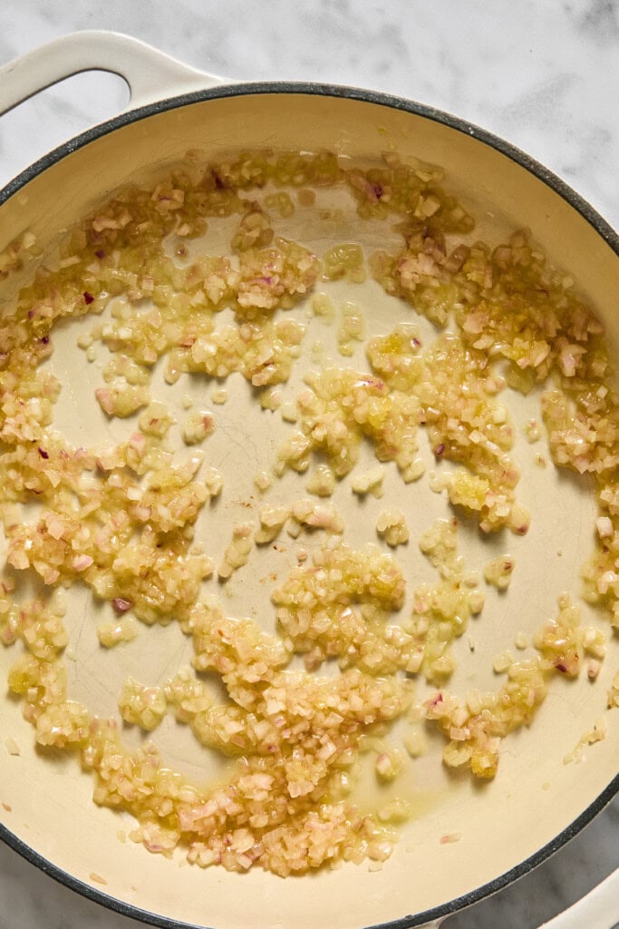 Chopped onions and garlic are sautéing in oil in a large cream-colored pot on a marble surface, forming the flavorful base for a creamy vegan pumpkin risotto.