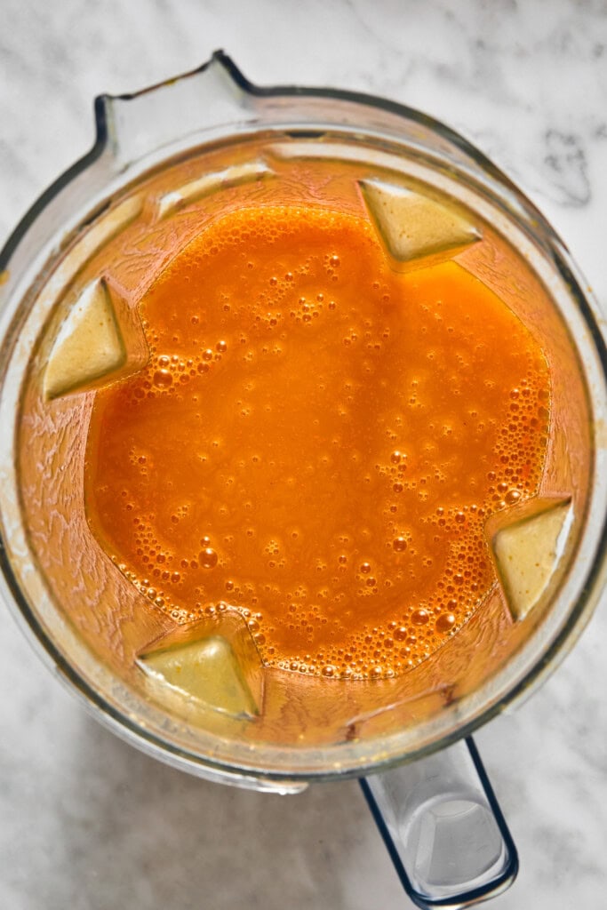 Overhead view of a blender filled with smooth, orange liquid on a marble surface. The mixture, perfect for vegan pumpkin risotto, appears freshly blended and slightly frothy, with the blender blades visible around the edges.