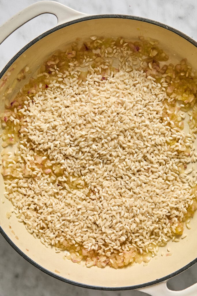 Uncooked risotto rice is added to a pot with sautéed onions and garlic, starting the vegan pumpkin risotto cooking process on a stovetop.