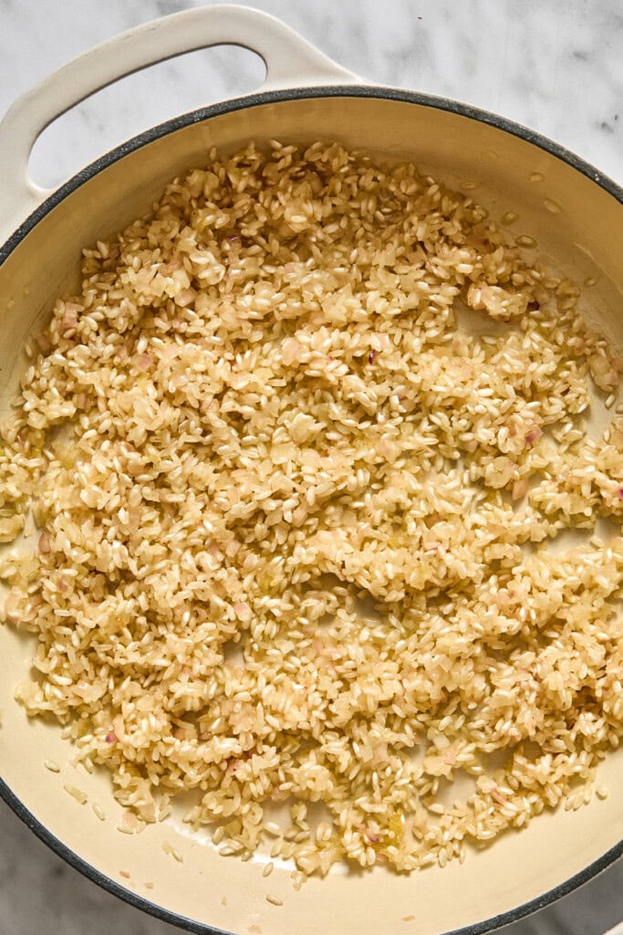 A close-up of a white pot on a marble surface, filled with uncooked arborio rice being sautéed, commonly the first step in making vegan pumpkin risotto.