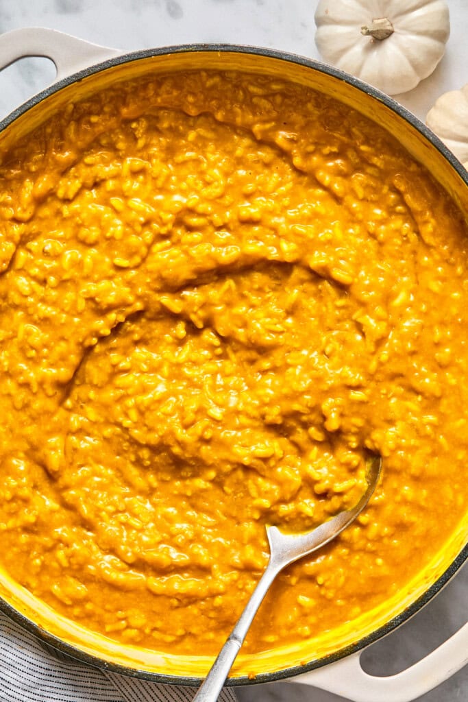 A large pot filled with creamy, orange-colored vegan pumpkin risotto, with a silver spoon resting inside. Small white pumpkins are visible in the background on a marble surface.