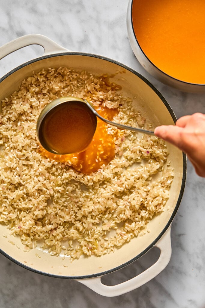 A hand pours orange broth from a ladle into a pan of sautéed rice, preparing vegan pumpkin risotto on a light marble countertop.