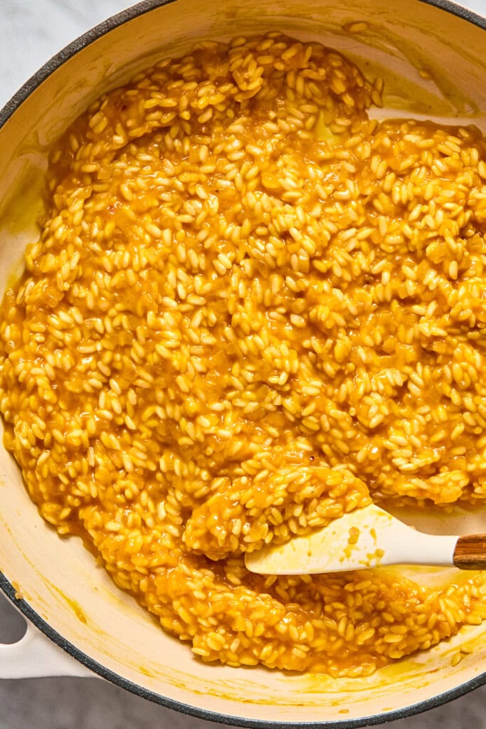 A pot filled with creamy, golden-yellow vegan pumpkin risotto being stirred with a white spatula.