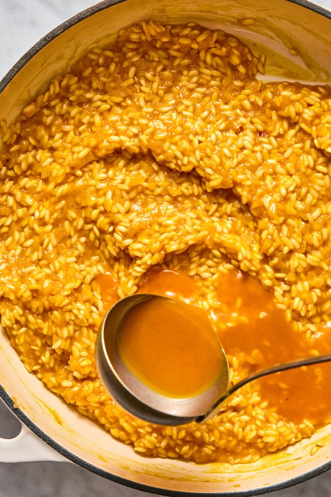 A pot of creamy vegan pumpkin risotto is shown, with a ladle scooping up some broth. The grains of rice appear tender and well-mixed with the rich sauce.