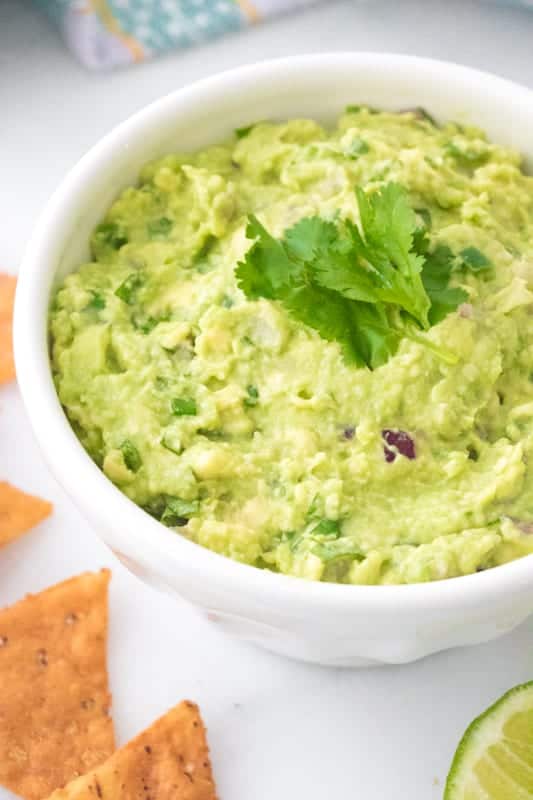 Guacamole served in a white bowl garnished with fresh cilantro and served with tortilla chips with a white background.