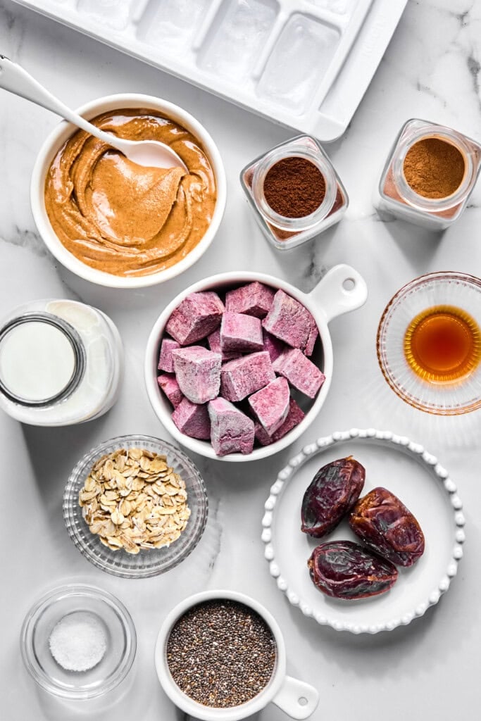 Top-down view of ingredients including frozen berries, dates, oats, chia seeds, nut butter, milk, spices, vanilla extract, salt, and an ice tray—all neatly arranged on a white surface—perfect for crafting a delicious purple sweet potato smoothie or experimenting with a nourishing no-fruit smoothie.