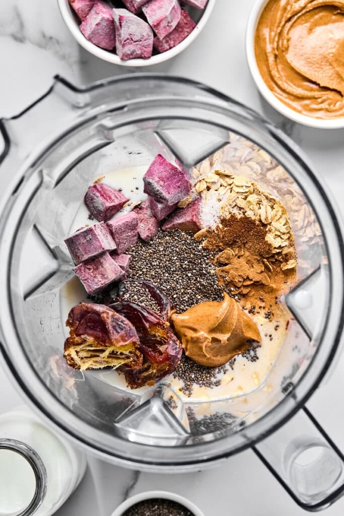 A blender contains frozen purple fruit, oats, chia seeds, dates, peanut butter, cinnamon, and milk. Surrounding the blender are bowls with more peanut butter, frozen fruit, chia seeds, and a bottle of milk—ideal ingredients to whip up a nourishing vegan sweet potato smoothie or even take inspiration for a delicious no-fruit smoothie variation.