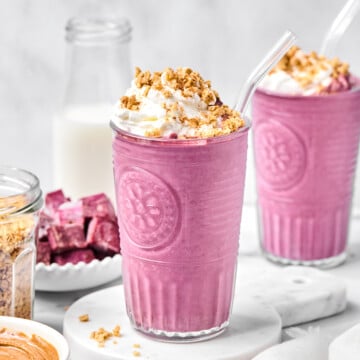 Two glasses of pink smoothie, possibly a vibrant purple sweet potato smoothie, are topped with whipped cream and granola, each with a clear straw. Surrounding items include a bottle of milk, a bowl of granola, a bowl of nut butter, and purple cubes on a plate.
