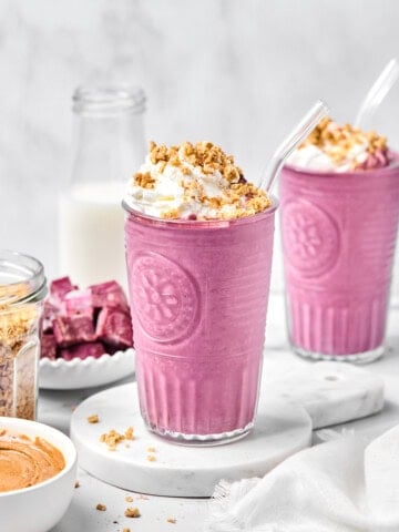 Two glasses of pink smoothie, possibly a vibrant purple sweet potato smoothie, are topped with whipped cream and granola, each with a clear straw. Surrounding items include a bottle of milk, a bowl of granola, a bowl of nut butter, and purple cubes on a plate.