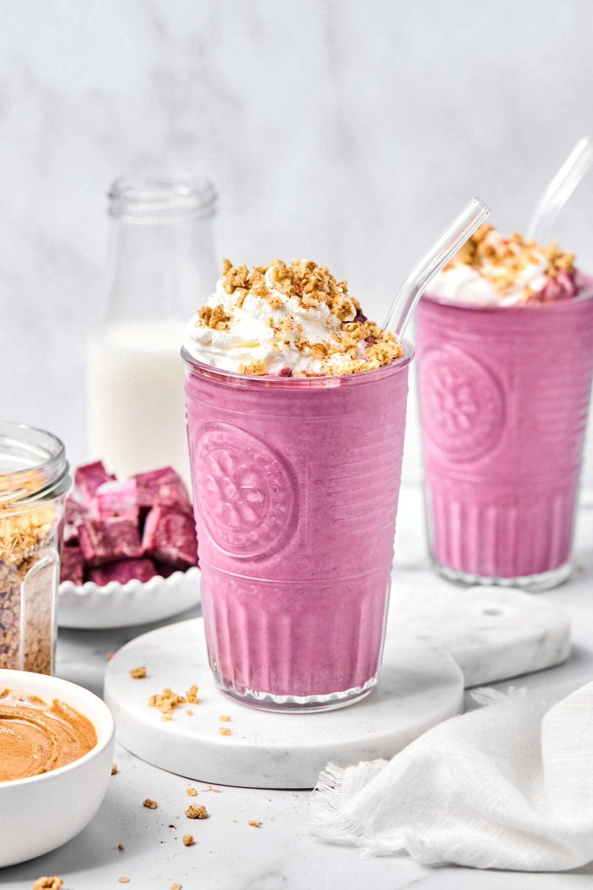 Two glasses of pink smoothie, possibly a vibrant purple sweet potato smoothie, are topped with whipped cream and granola, each with a clear straw. Surrounding items include a bottle of milk, a bowl of granola, a bowl of nut butter, and purple cubes on a plate.