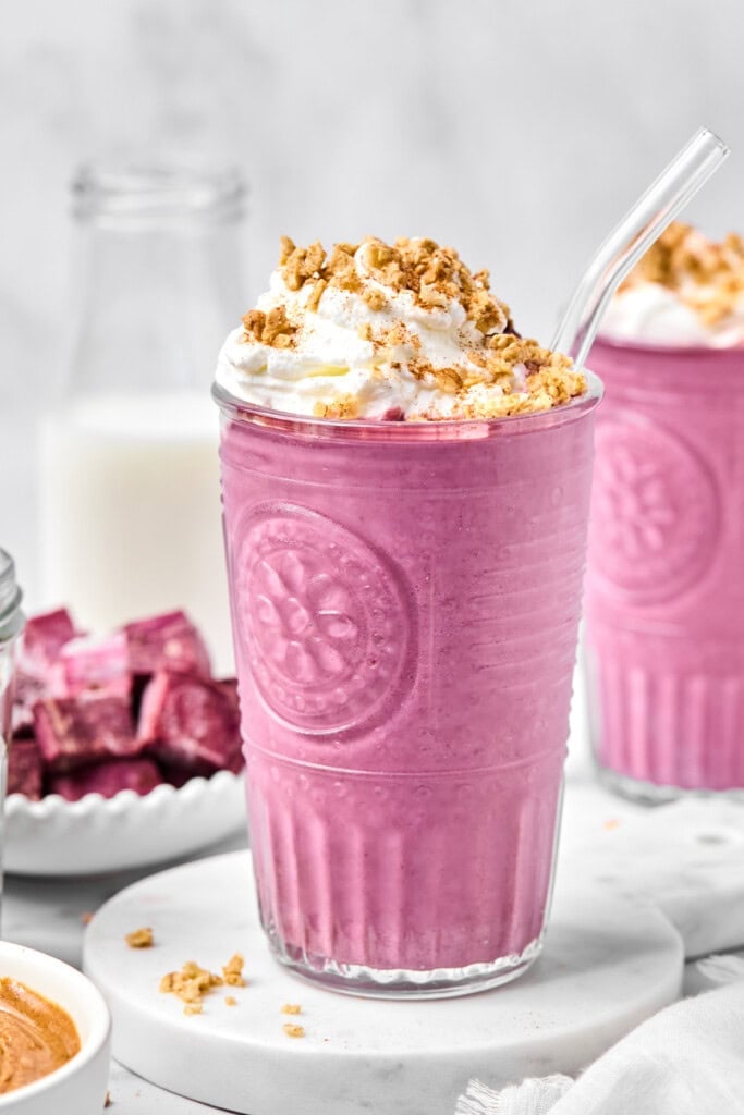 A creamy pink, fruit-free smoothie topped with whipped cream and granola sits in a clear glass with a glass straw. In the background, there’s a bottle of milk and a plate with purple cubes.