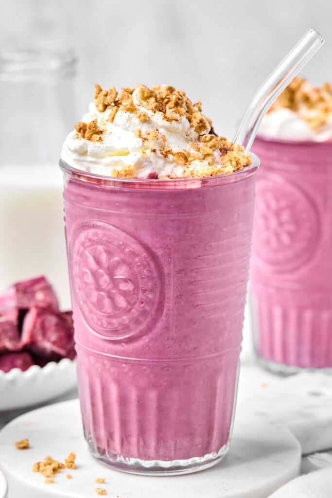 A vibrant pink vegan sweet potato smoothie topped with whipped cream and granola sits in a textured glass with a glass straw. Another similar no-fruit smoothie and a bottle of milk are blurred in the background.
