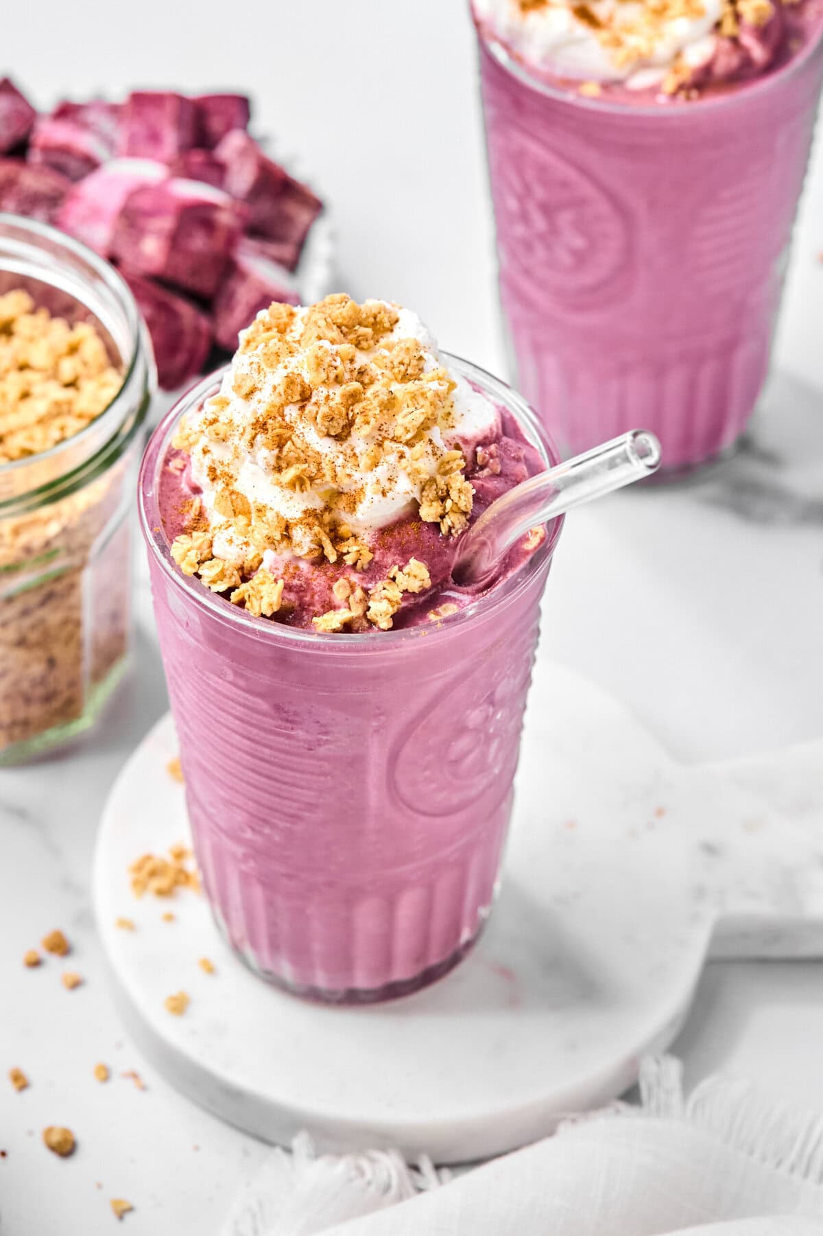 A pink no-fruit smoothie in a clear glass with a glass straw, topped with whipped cream and granola. There’s a jar of granola and another smoothie in the background on a white surface.
