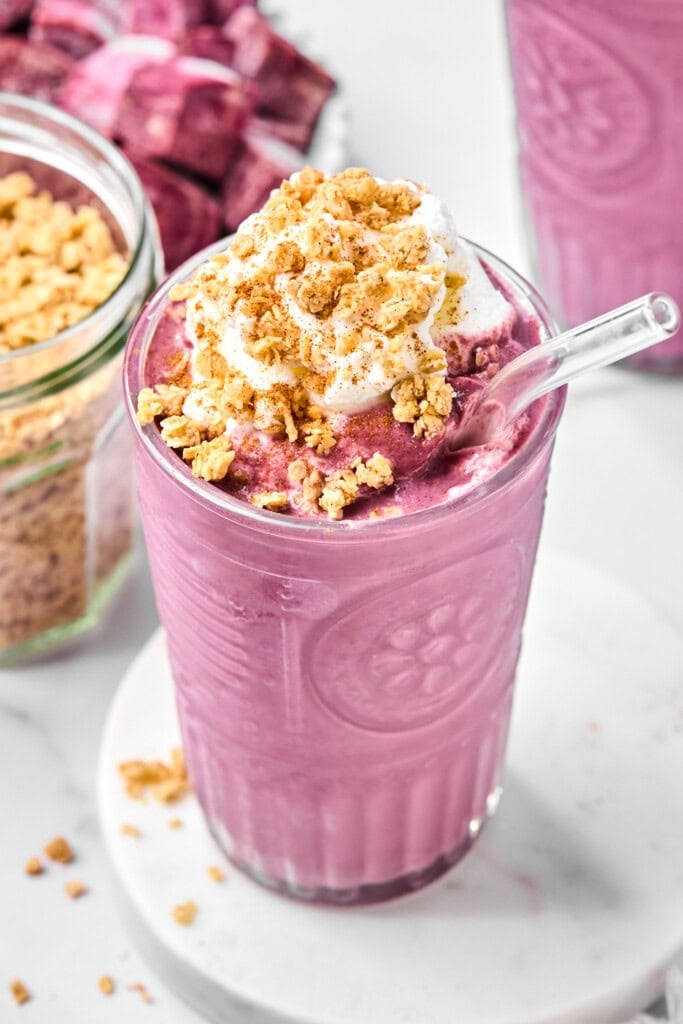 A vibrant vegan sweet potato smoothie, topped with fluffy whipped cream, crunchy granola, and a dusting of cinnamon, is served in a clear glass with a glass straw. Granola and other smoothie ingredients are visible in the background.