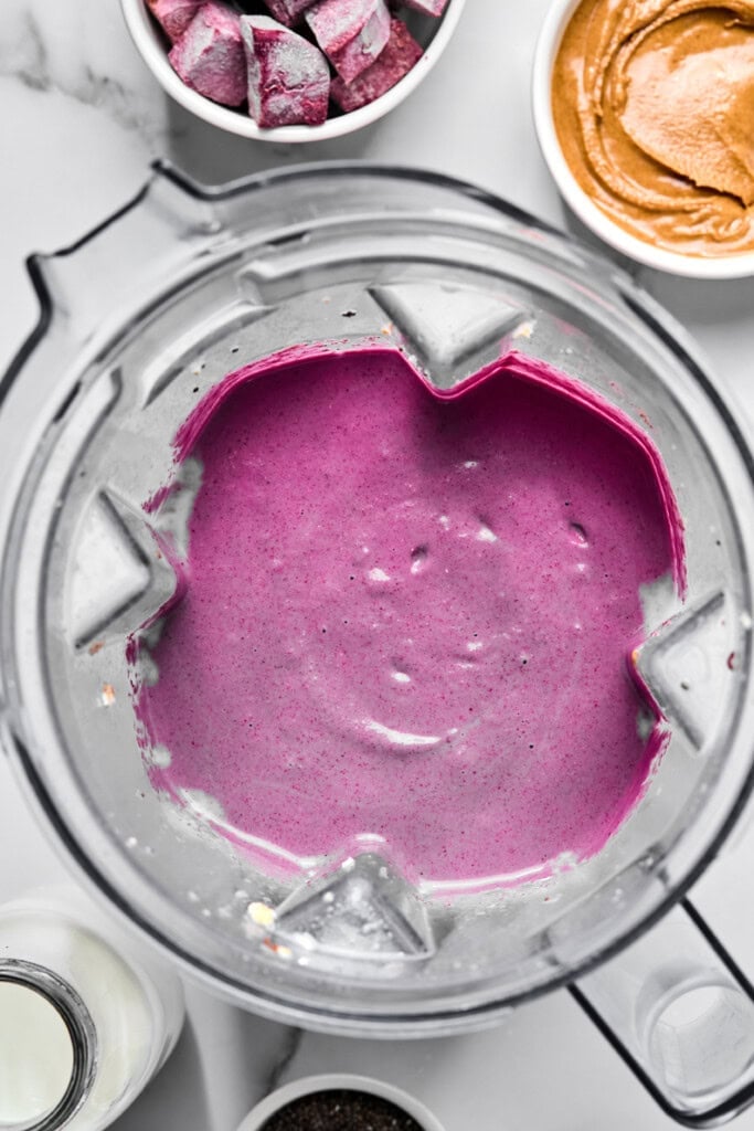 A blender filled with a smooth, vibrant purple vegan sweet potato smoothie is surrounded by bowls of frozen berries, peanut butter, chia seeds, and a bottle of milk on a white surface.