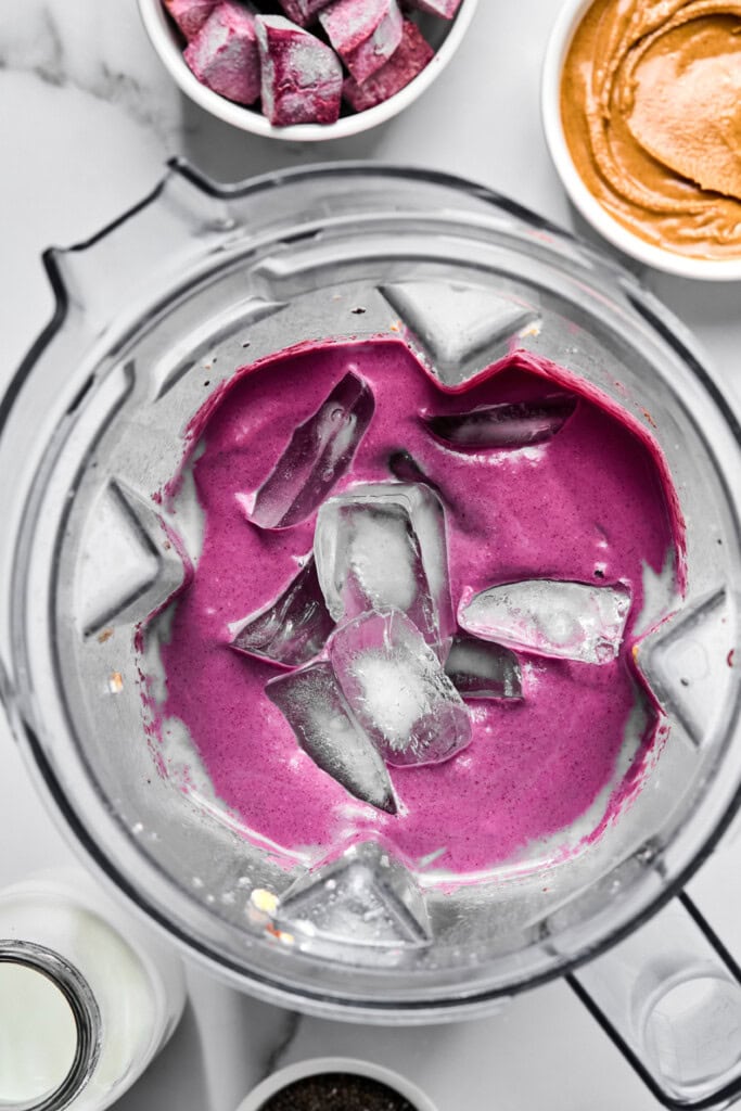 A blender filled with a vibrant purple sweet potato smoothie mixture and several ice cubes, surrounded by bowls of nut butter, frozen fruit, and a bottle of milk.