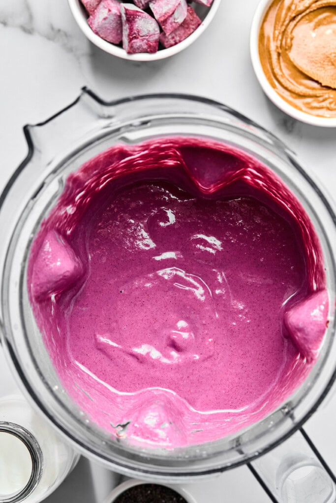 A blender filled with a thick, vibrant purple fruitless smoothie is surrounded by bowls of peanut butter, purple vegetable chunks, and a glass of milk on a white surface.