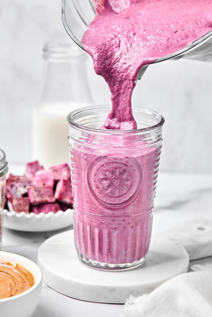 A thick purple sweet potato smoothie is being poured into a clear glass. Surrounding the glass are a bowl of nut butter, a bowl of purple fruit pieces, and a bottle of milk, all on a white surface.
