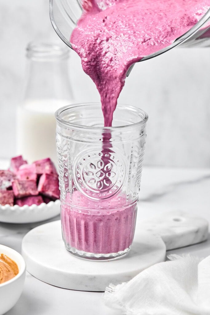 A thick, vibrant pink vegetable smoothie is being poured from a blender into a clear glass. There are beets, a bowl of peanut butter, and a milk bottle in the background on a white surface—perfect ingredients for whipping up a creamy vegan sweet potato smoothie.