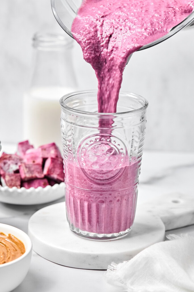 Bright pink fruit-free smoothie being poured from a blender into a clear glass on a white surface, with a bowl of pink cubes, a bowl of nut butter, and a bottle of milk in the background.