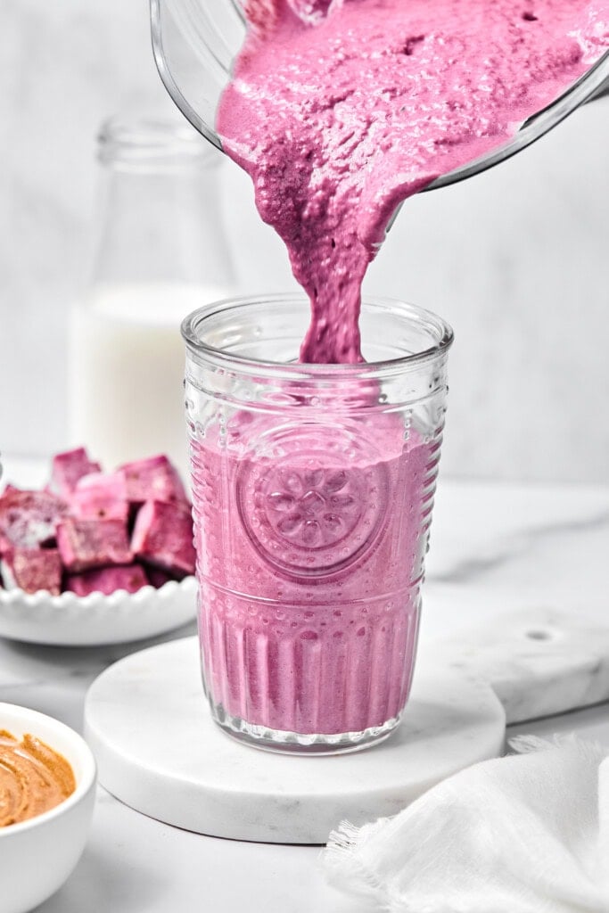 A thick, bright pink smoothie is being poured from a blender into a clear glass, with a bowl of chopped fruit, a bowl of nut butter, and a bottle of milk in the background—perfect for anyone craving a flavorful fruitless smoothie or looking to try the vibrant twist of a purple sweet potato smoothie.