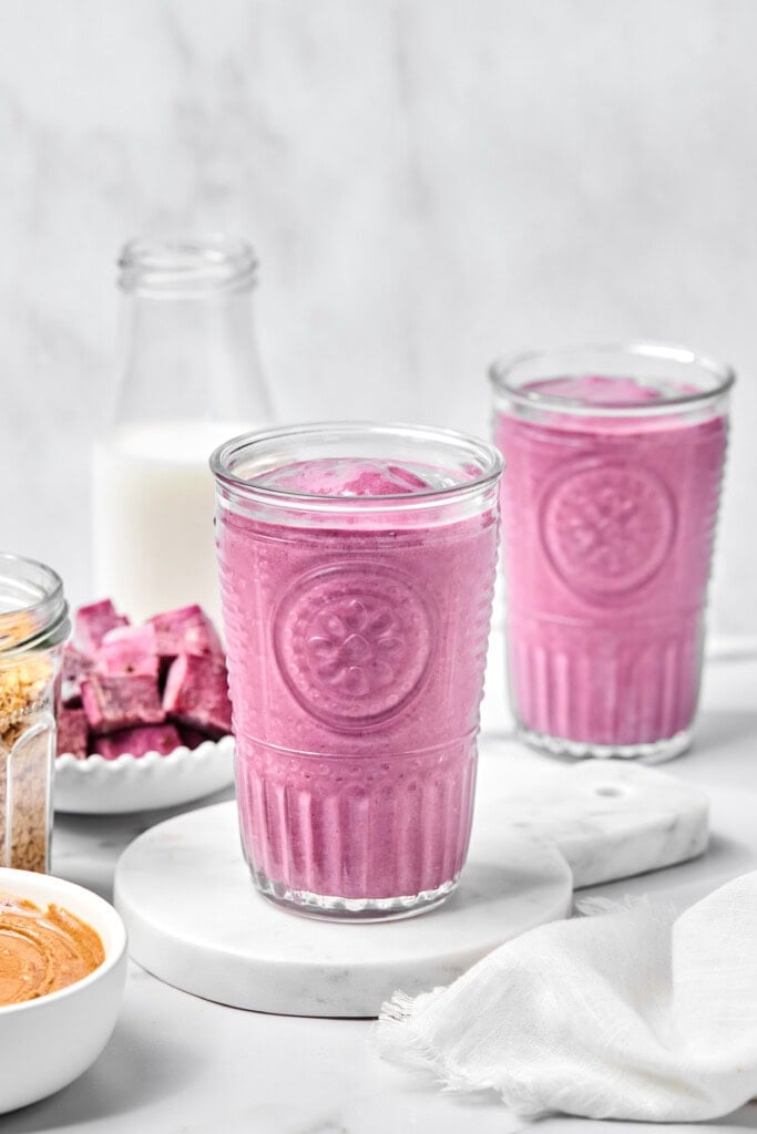 Two clear glasses filled with pink smoothie sit on white coasters. Behind them are a glass bottle of milk, a bowl of nut butter, a jar of granola, pink fruit pieces, and a white napkin on a light surface—setting the scene for this creamy vegan sweet potato smoothie that's both vibrant and nourishing.