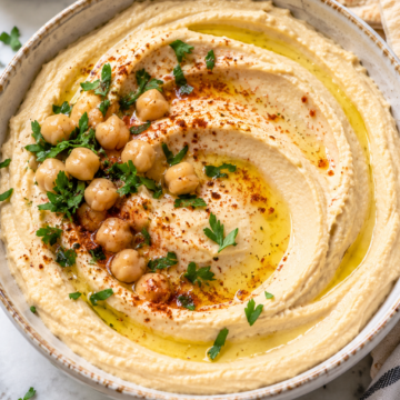 A bowl of creamy Mediterranean hummus topped with whole chickpeas, olive oil, paprika, and chopped parsley, with pieces of pita bread and a small bowl of herbs on the side.
