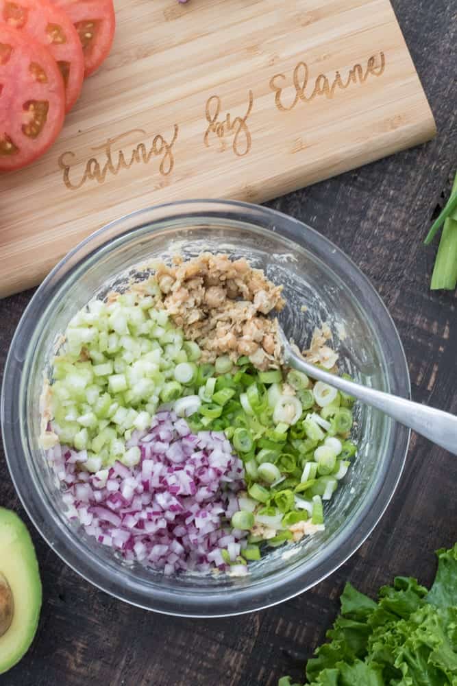 Vegan chickpea salad ingredients in a glass mixing bowl prior to being tossed together next to a cutting board.