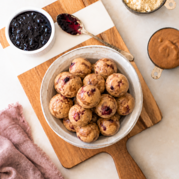 A bowl of round almond butter jelly energy bites with visible berry bits sits on a wooden board, surrounded by bowls of jam, almond flour, and nut butter, with a spoon and a mauve cloth nearby.