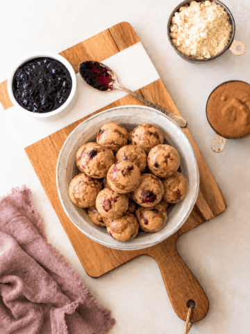 A bowl of round almond butter jelly energy bites with visible berry bits sits on a wooden board, surrounded by bowls of jam, almond flour, and nut butter, with a spoon and a mauve cloth nearby.