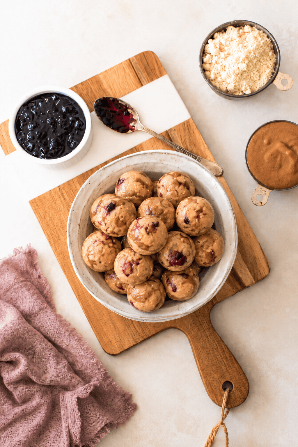 A bowl of round almond butter jelly energy bites with visible berry bits sits on a wooden board, surrounded by bowls of jam, almond flour, and nut butter, with a spoon and a mauve cloth nearby.