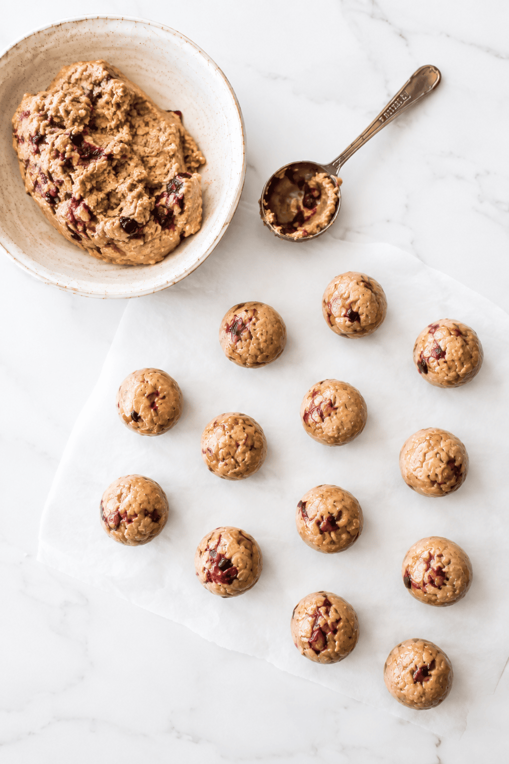 Overhead view of a bowl of oatmeal cookie dough with cranberries, reminiscent of almond butter jelly energy bites, a spoon, and fourteen round dough balls arranged on parchment paper on a marble surface.