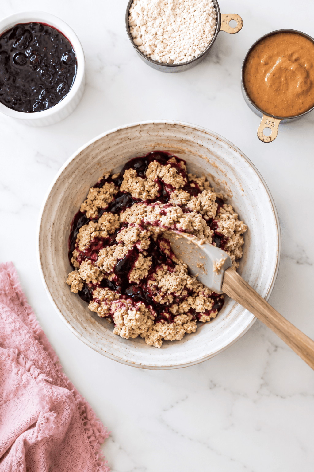 A mixing bowl with oat crumble and berry filling, reminiscent of almond butter jelly energy bites, is surrounded by a cup of flour, a cup of nut butter, a bowl of berry jam, and a pink cloth on a white marble surface.