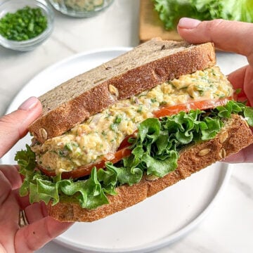 A person holds a sandwich filled with leafy lettuce, sliced tomato, and a creamy vegan chickpea avocado egg salad between two slices of whole grain bread. Fresh vegetables and a jar of salad spread are visible in the background.