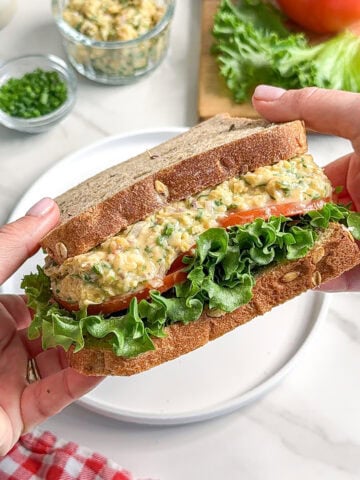 A person holds a sandwich filled with leafy lettuce, sliced tomato, and a creamy vegan chickpea avocado egg salad between two slices of whole grain bread. Fresh vegetables and a jar of salad spread are visible in the background.