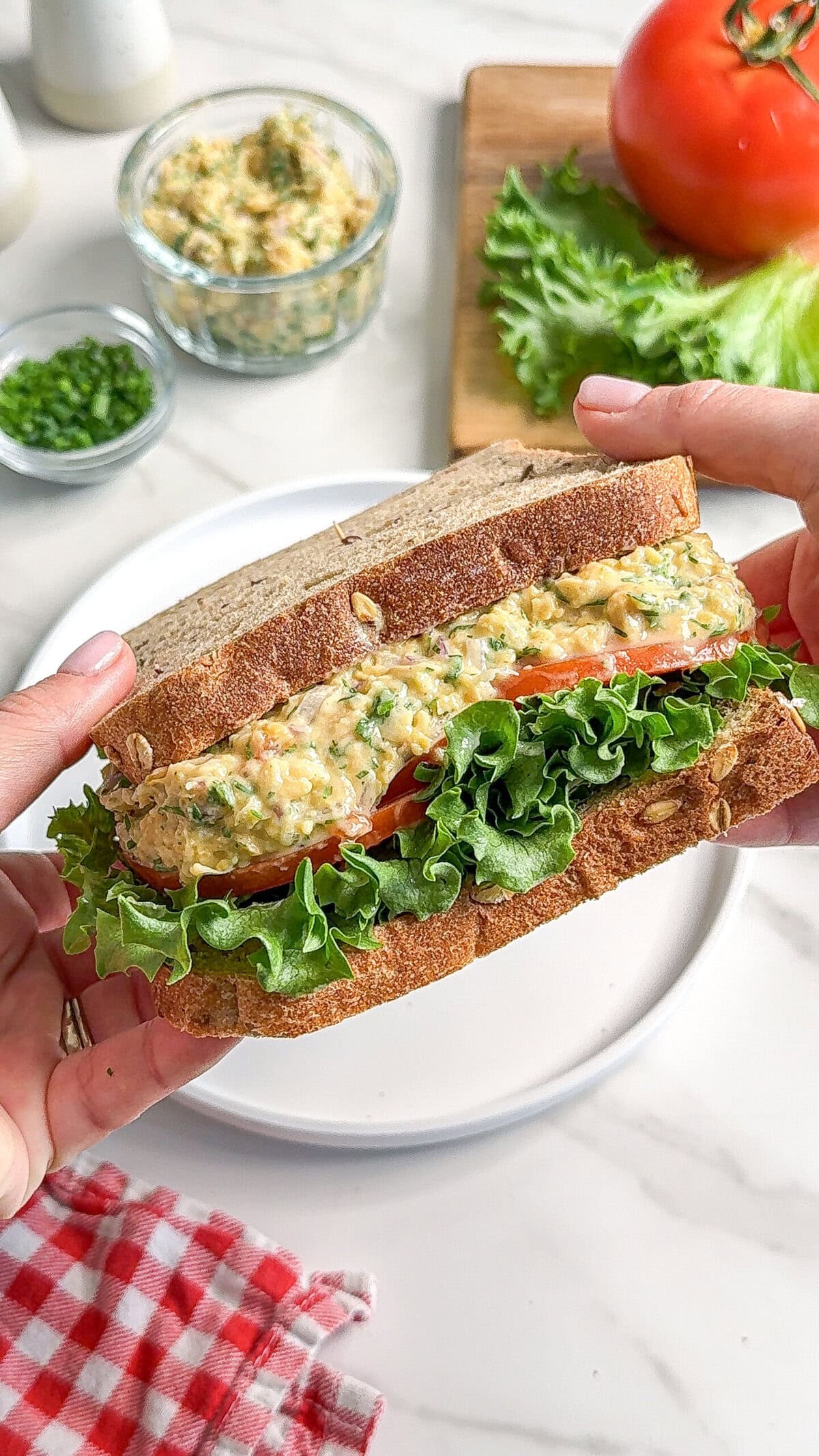 A person holds a sandwich filled with leafy lettuce, sliced tomato, and a creamy vegan chickpea avocado egg salad between two slices of whole grain bread. Fresh vegetables and a jar of salad spread are visible in the background.