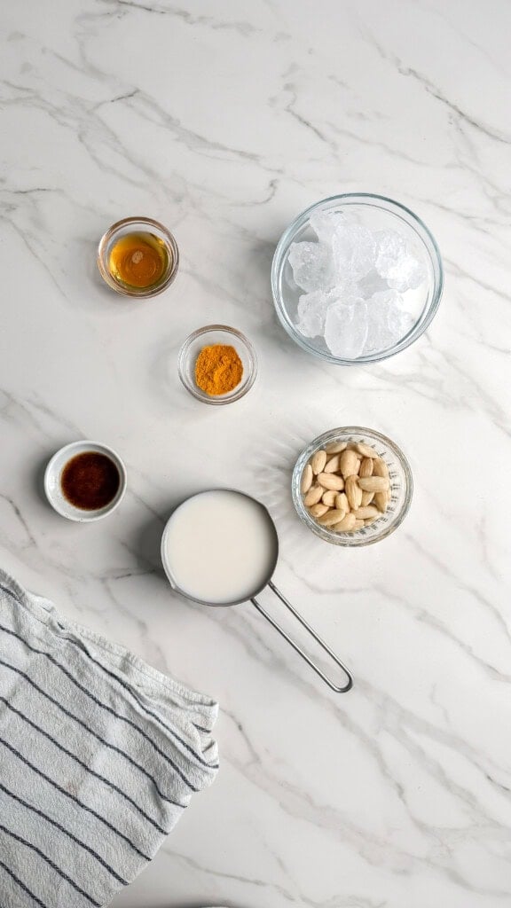 A flat lay on marble: a bowl of ice, blanched almonds, milk, ground turmeric, vanilla extract, honey, and a striped towel—everything you need to create a refreshing vegan iced turmeric latte at home.