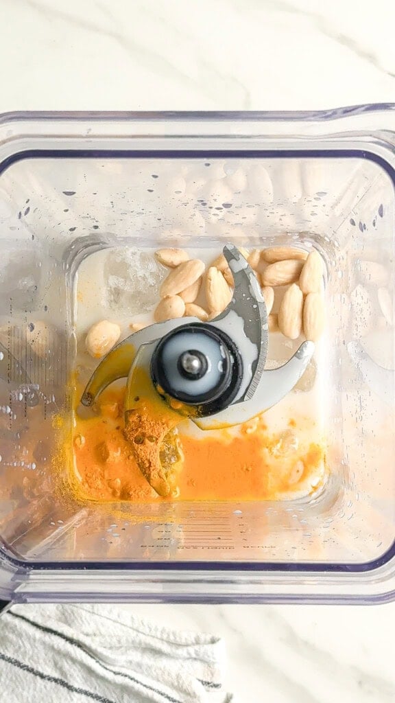 A top-down view of a blender with blanched almonds, turmeric powder, and liquid ingredients inside, ready to be blended for a creamy vegan iced turmeric latte. The blender sits on a light-colored countertop next to a striped cloth.