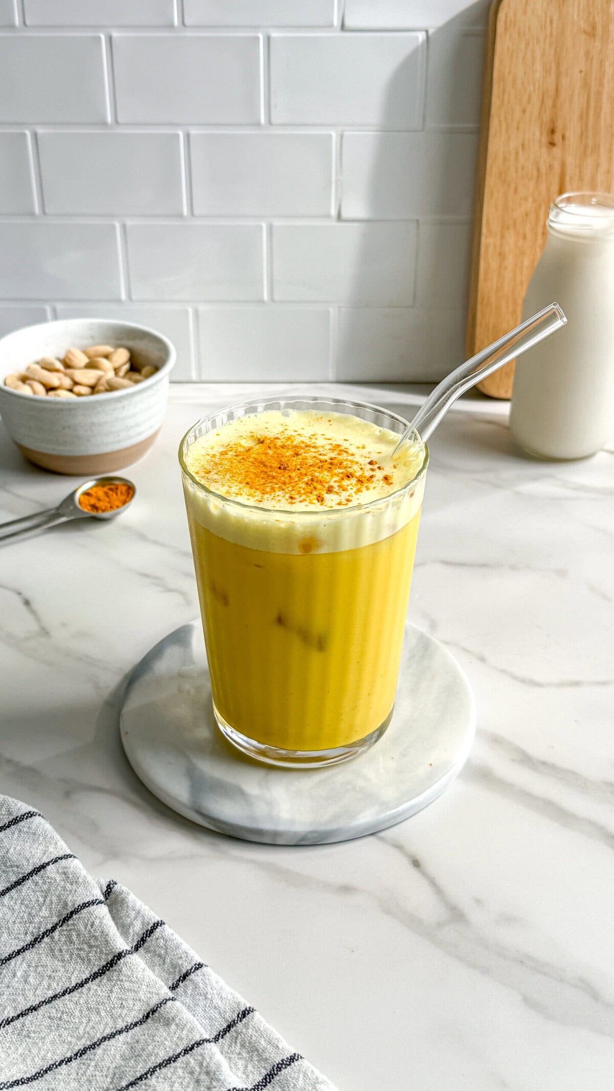 A glass of vegan iced turmeric latte with a glass straw sits on a marble coaster. A bowl of almonds, a spoon with yellow spice, a jar of milk, and a wooden board are in the background on a white countertop.