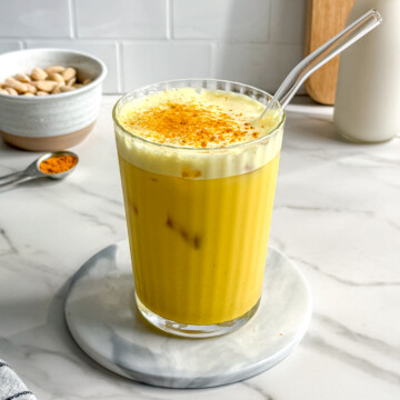 A glass of golden yellow vegan iced turmeric latte with a glass straw sits on a marble coaster. In the background are a bowl of pistachios, a spoon of turmeric, and a glass bottle of milk on a white countertop.