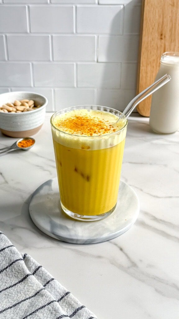 A glass of golden yellow vegan iced turmeric latte with a glass straw sits on a marble coaster. In the background are a bowl of pistachios, a spoon of turmeric, and a glass bottle of milk on a white countertop.