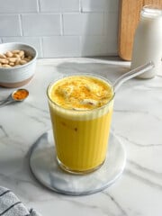 A glass of golden yellow vegan iced turmeric latte with a glass straw sits on a round marble coaster. In the background are a bowl of almonds, a spoon with turmeric powder, and a bottle of milk on a marble countertop.